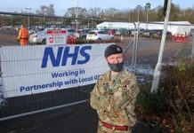 Reservist at heart of the North’s mass vaccination effort – Case study Man in British Army uniform standing with his arms crossed in front of a sign that reads NHS.